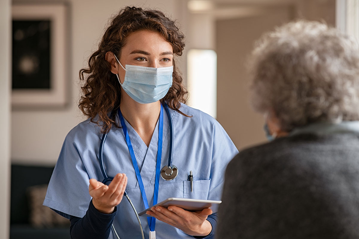 A masked female health care worker speaks with an older female patient.