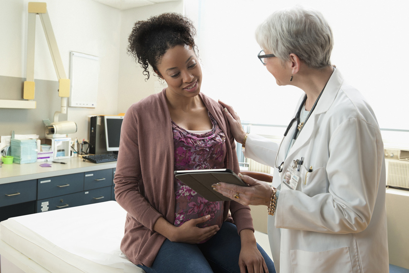 A pregnant woman cradles her belly while looking at a tablet held by her doctor.