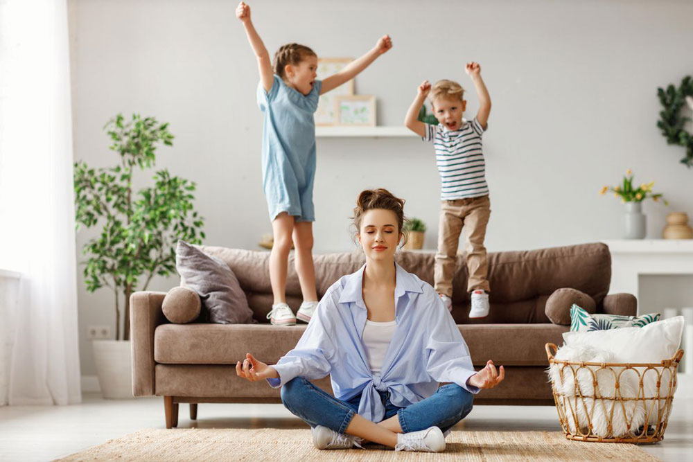 mom meditates on floor while kids jump on sofa