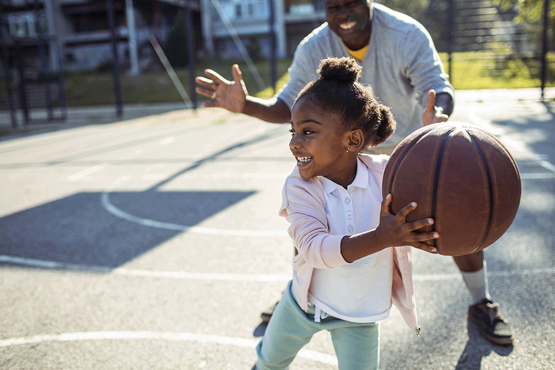 A young girl smiles as she plays basketball with her father.