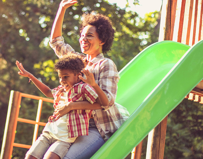 A mother and child smile as they play on a slide at a park.