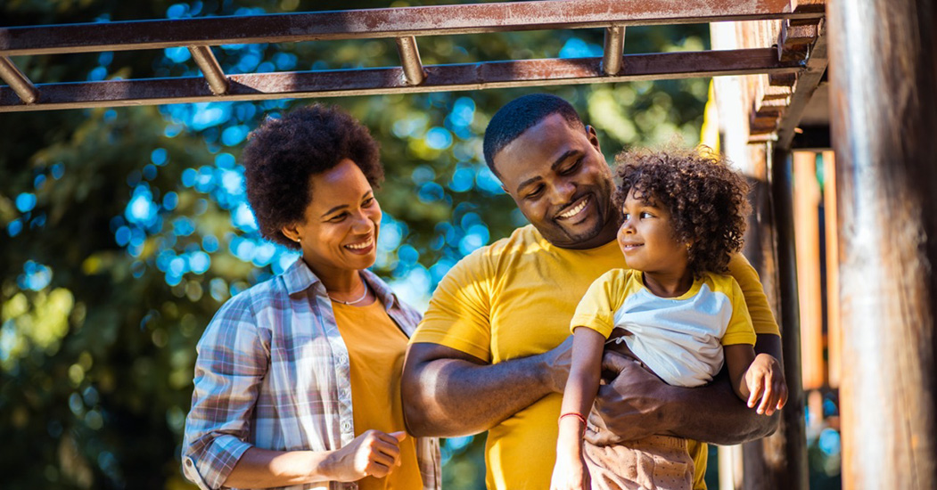 A mother, father and young child play near a jungle gym at a park.