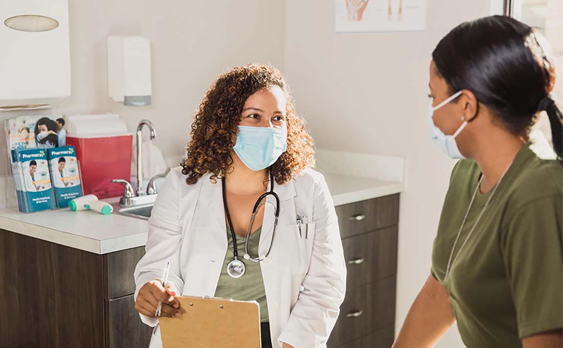 A female physician wears a mask and holds a clipboard while speaking with a young female patient.