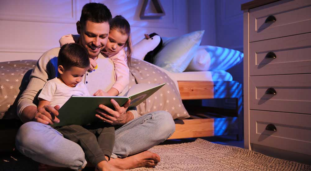 father sitting beside bed with two children reading a book